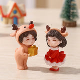Plastic Christmas figurines in peach and red stand on pale wood, with a miniature house and tree blurred behind them.