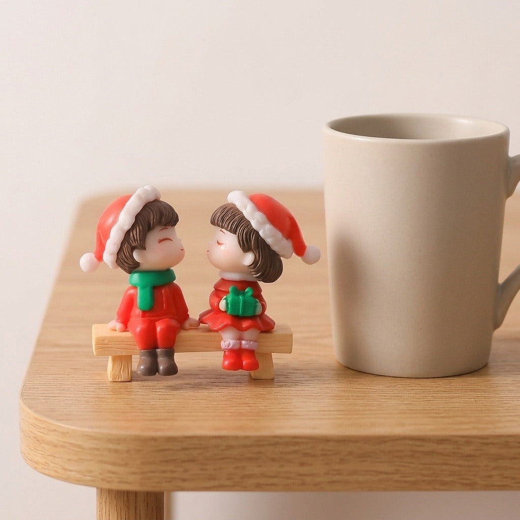 Two plastic Christmas figurines wearing red Santa hats sit on a miniature tan bench beside a beige mug on a wooden table.
