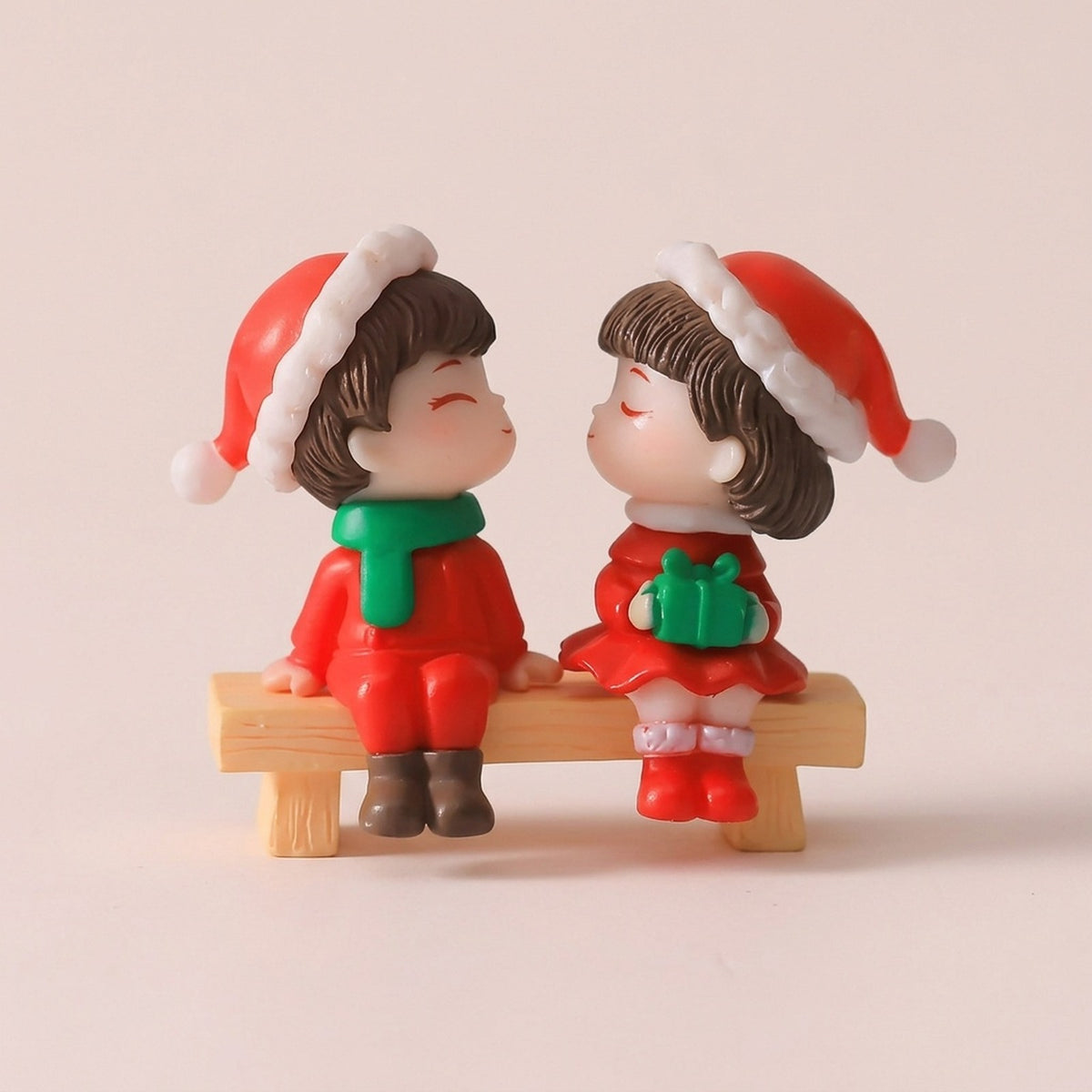Two plastic Christmas figurines in red Santa hats sit on a tan bench against beige, with a green scarf and green gift.