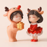 Plastic Christmas figurines stand against beige, one wearing a tan reindeer suit and holding a gold gift, the other wearing a red antlered dress.