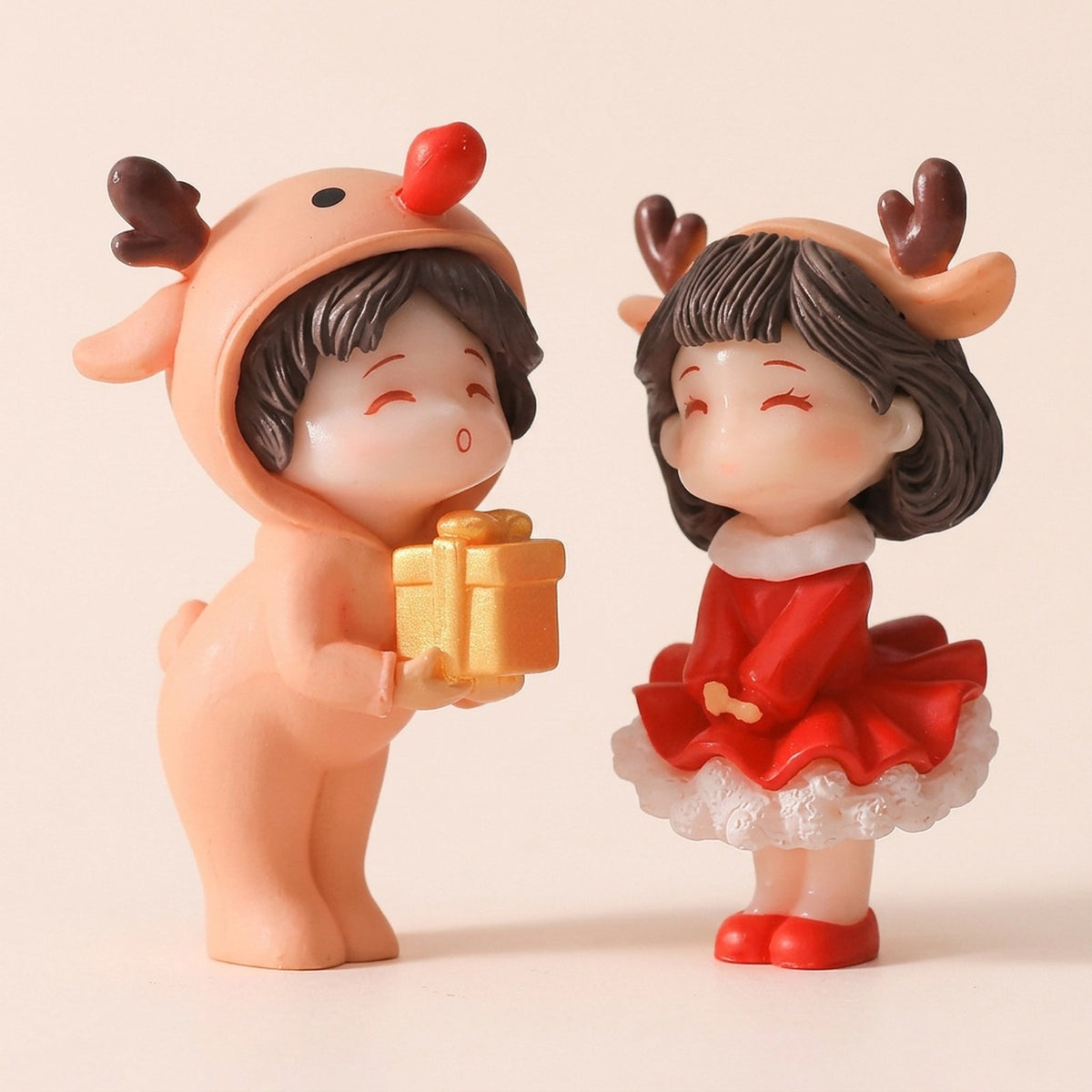 Plastic Christmas figurines stand against beige, one wearing a tan reindeer suit and holding a gold gift, the other wearing a red antlered dress.