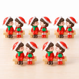 Five pairs of plastic Christmas figurines in red outfits and Santa hats sit on tan benches in two rows on a pale surface.