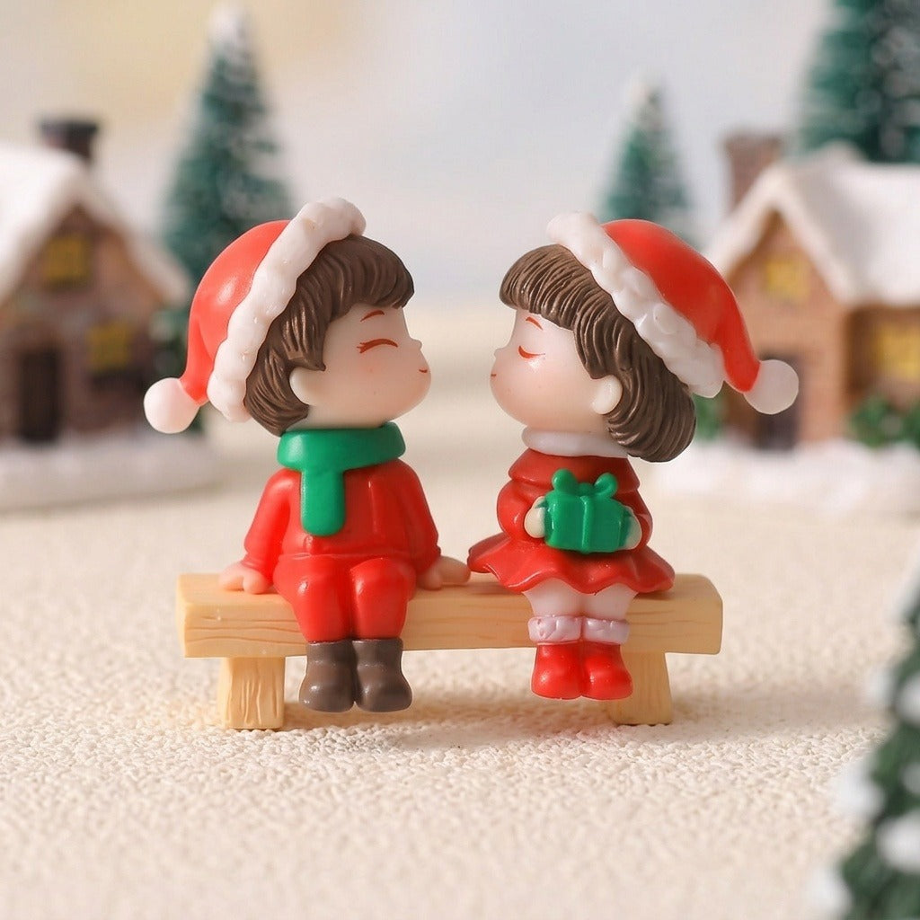 Plastic Christmas figurines wearing red Santa hats sit together on a tan bench among miniature snowy houses and green trees.