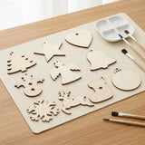 Pale wooden ornament blanks in mixed Christmas shapes lie on a beige mat beside paintbrushes and a white palette.