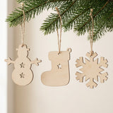 Pale wooden ornament blanks shaped as a snowman, stocking, and snowflake hang by brown twine from evergreen branches against a cream background.
