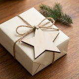A pale wooden star ornament blank rests on a cream-wrapped gift tied with brown twine beside evergreen foliage on a wooden table.