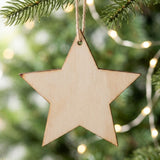 A pale wooden star ornament blank with dark edges hangs by brown twine from an evergreen branch against blurred warm lights.