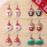 Six pairs of colorful Christmas drop earrings with silver hooks, including snowmen, penguins, Santas, and trees, arranged on light wood.
