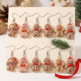 Twelve tan gingerbread drop earrings with silver hooks are displayed on a cream card beside an evergreen sprig and red ribbon.