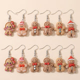 Twelve tan gingerbread drop earrings with red bows, Santa hats, and contrasting buttons lie in two rows on a cream surface.