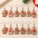 Twelve tan gingerbread-character drop earrings with silver fishhooks lie in two rows on a cream surface beside a fir sprig and red ribbon.
