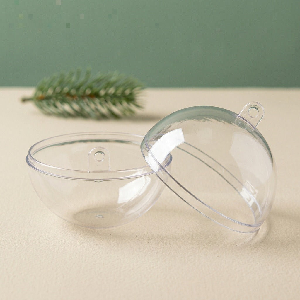 A clear plastic fillable ornament rests in two separated halves on a cream surface beside an evergreen sprig against a green background.