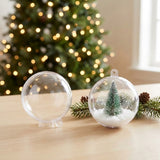 Two clear plastic fillable ornaments sit on a wooden table, one empty and one holding a miniature green tree and artificial snow.