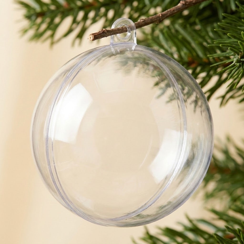 An empty clear plastic fillable ornament hangs from an evergreen twig through its top tab against a beige background.