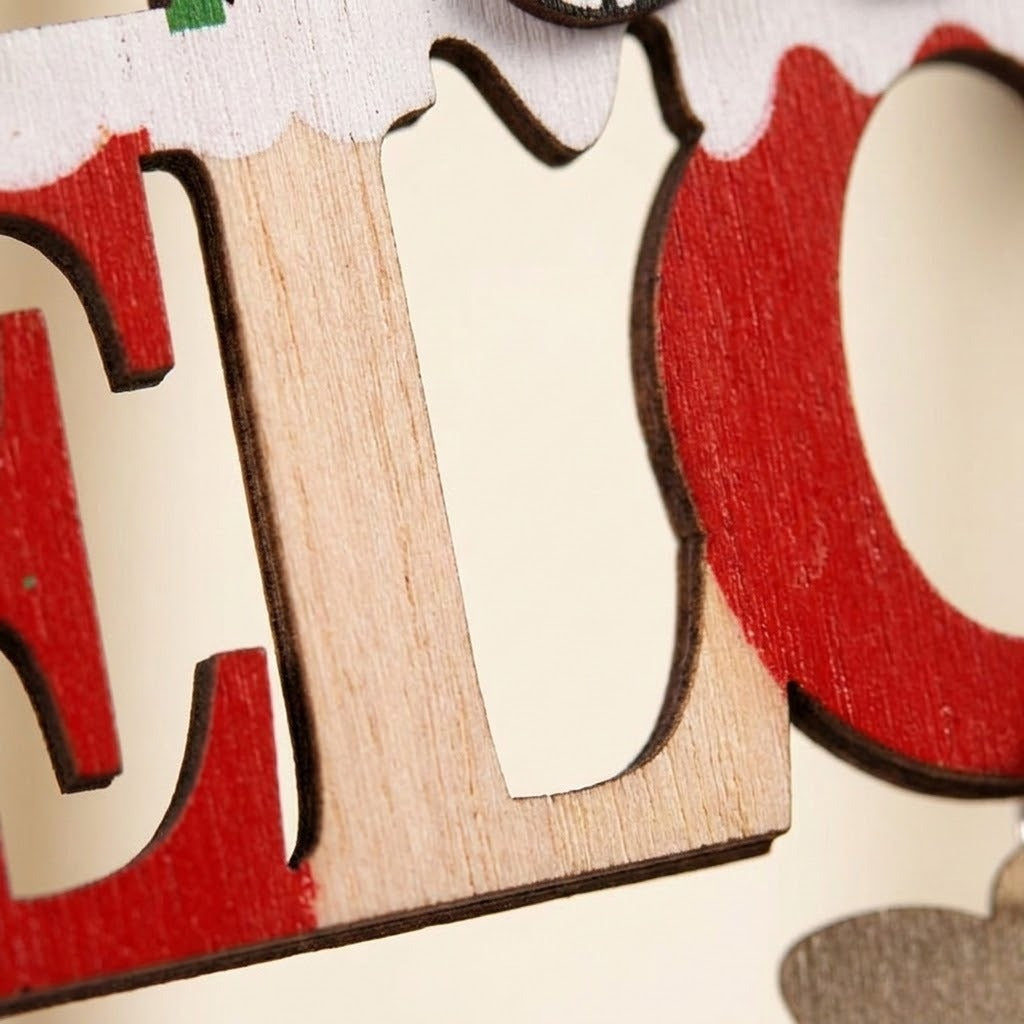 Close-up of the wooden Christmas ornament shows natural wood grain, red painted letters, white snow detailing, and dark edges against cream.