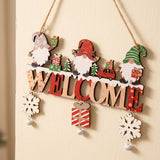 Red and natural wooden Christmas ornament with gnomes, WELCOME letters, snowflakes, and bells hangs at an angle against a cream wall.
