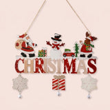 Wooden Christmas ornament with Santa, snowman, reindeer, red and natural lettering, dangling snowflakes, a striped gift, and bells hangs against pale pink.