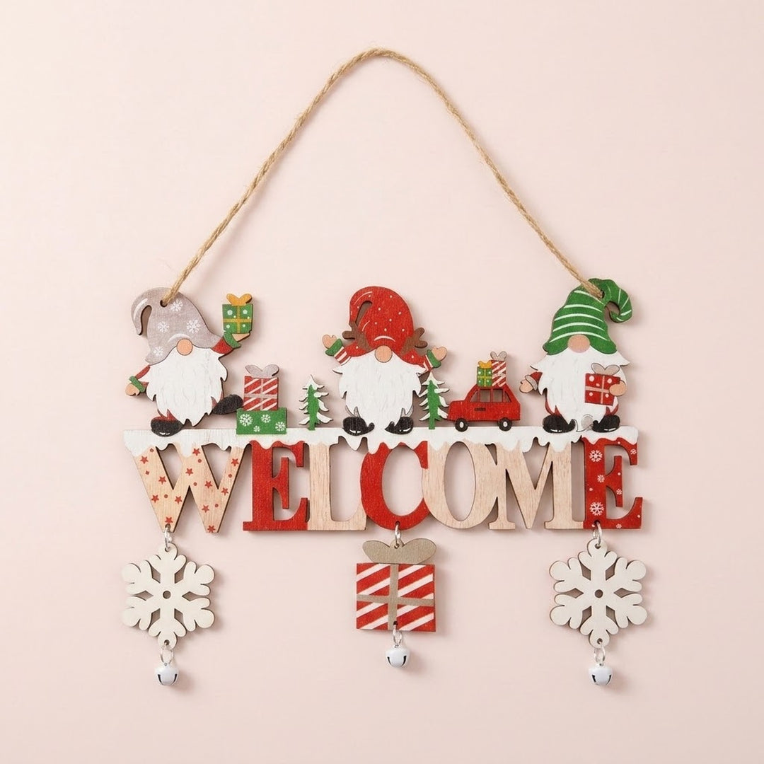 Wooden Christmas ornament with three gnomes, red and natural WELCOME lettering, dangling snowflakes, a striped gift, and bells hangs against pale pink.