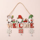 Wooden Christmas ornament with three gnomes, red and natural WELCOME lettering, dangling snowflakes, a striped gift, and bells hangs against pale pink.