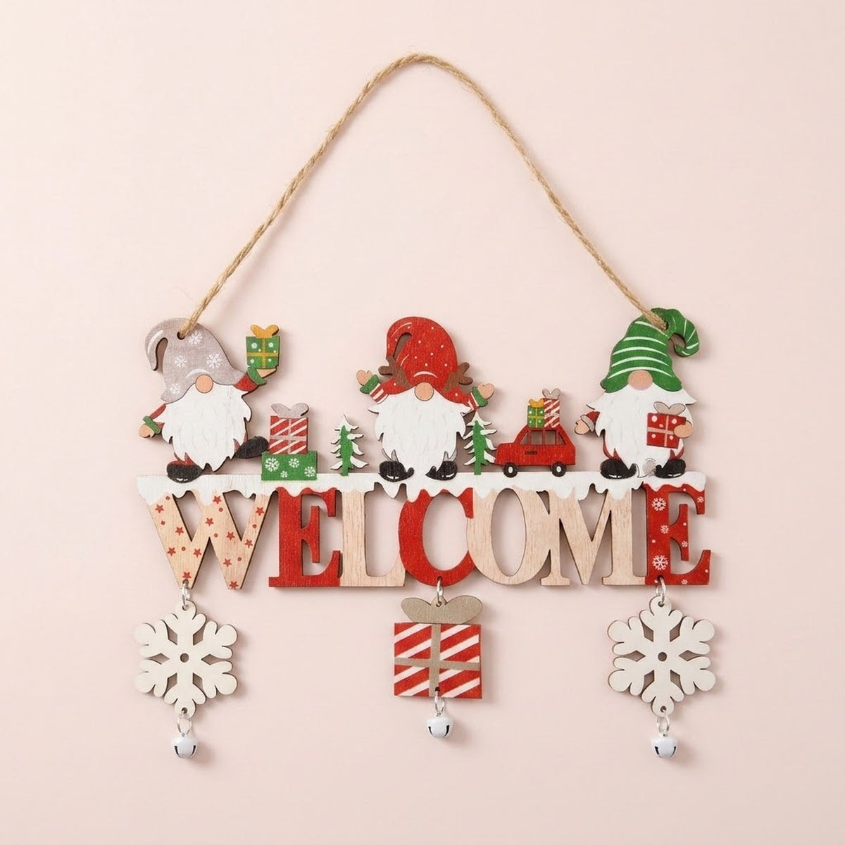 Wooden Christmas ornament with three gnomes, red and natural WELCOME lettering, dangling snowflakes, a striped gift, and bells hangs against pale pink.