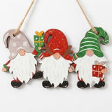 Wooden Christmas ornament features three white-bearded gnomes wearing gray, red, and green hats, suspended by twine against a white background.