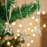 Three clear acrylic Christmas ornaments shaped like five-point stars hang from white ribbons on green branches against warm blurred lights.