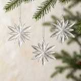 Three clear acrylic Christmas ornaments with faceted starburst shapes hang from silver cords beneath evergreen branches against a cream background.