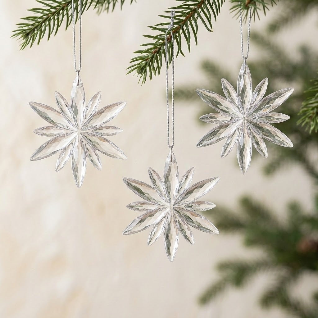 Three clear acrylic Christmas ornaments with faceted starburst shapes hang from silver cords beneath evergreen branches against a cream background.