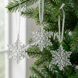 Three clear acrylic Christmas ornaments shaped like six-armed snowflakes hang from white ribbons on evergreen branches beside a bright window.