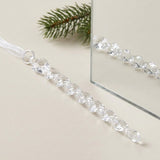 A clear acrylic twisted icicle Christmas ornament with a white ribbon rests beside a mirror and evergreen sprig on a cream surface.