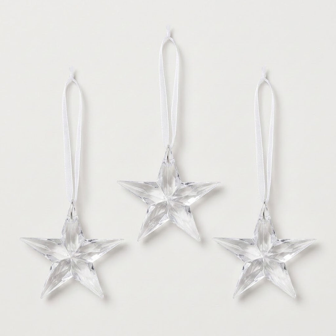 Three clear acrylic five-point star Christmas ornaments with faceted surfaces and white hanging ribbons are arranged against a white background.