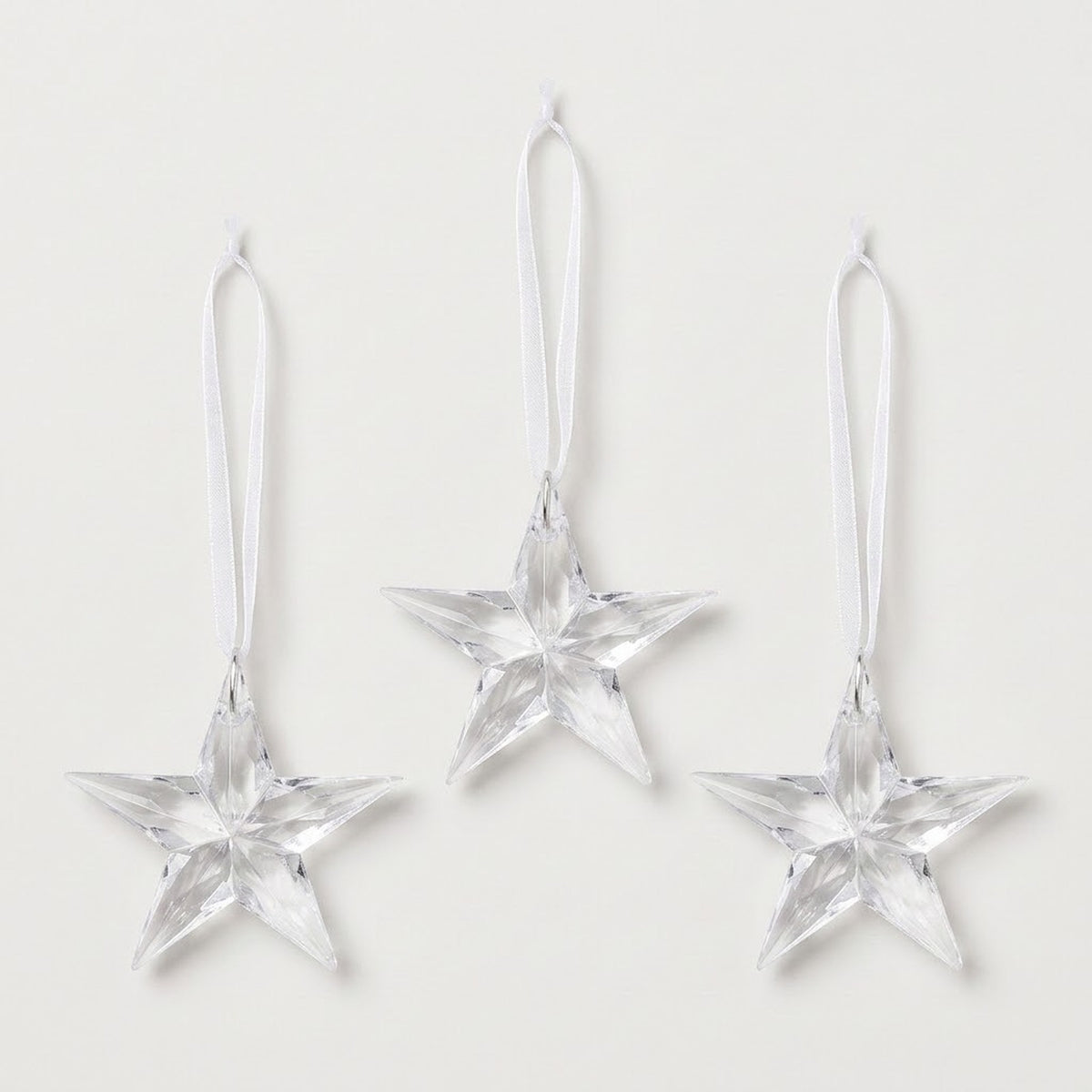 Three clear acrylic five-point star Christmas ornaments with faceted surfaces and white hanging ribbons are arranged against a white background.