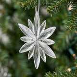 A clear acrylic Christmas ornament with a faceted starburst shape hangs from a white ribbon among green branches against softly blurred lights.