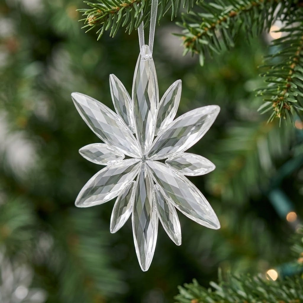 A clear acrylic Christmas ornament with a faceted starburst shape hangs from a white ribbon among green branches against softly blurred lights.
