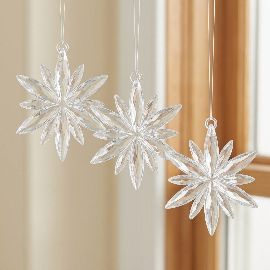 Three clear acrylic starburst Christmas ornaments hang at staggered heights beside a wooden window frame and cream curtain.