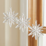 Three clear acrylic starburst Christmas ornaments hang at staggered heights beside a wooden window frame and cream curtain.