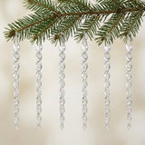 Six clear acrylic twisted icicle Christmas ornaments hang from a green evergreen branch against a softly blurred cream background.