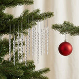 Clear twisted icicle ornaments hang from green Christmas tree branches beside a red bauble against cream curtains.