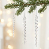 Three clear twisted icicle ornaments hang from silver cords beneath an evergreen branch against a pale background with warm blurred lights.