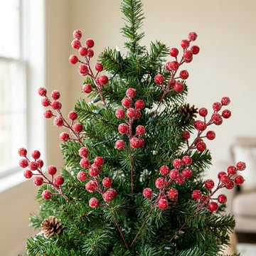 Frosted red berry picks decorate a green Christmas tree with pinecones beside a window in a beige room.