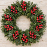 Frosted red berry picks are arranged around a green evergreen wreath on pale wooden planks.
