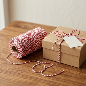 A roll of red and white striped bakers twine sits on a wooden table next to a brown kraft gift box tied with the same twine and a blank paper tag.