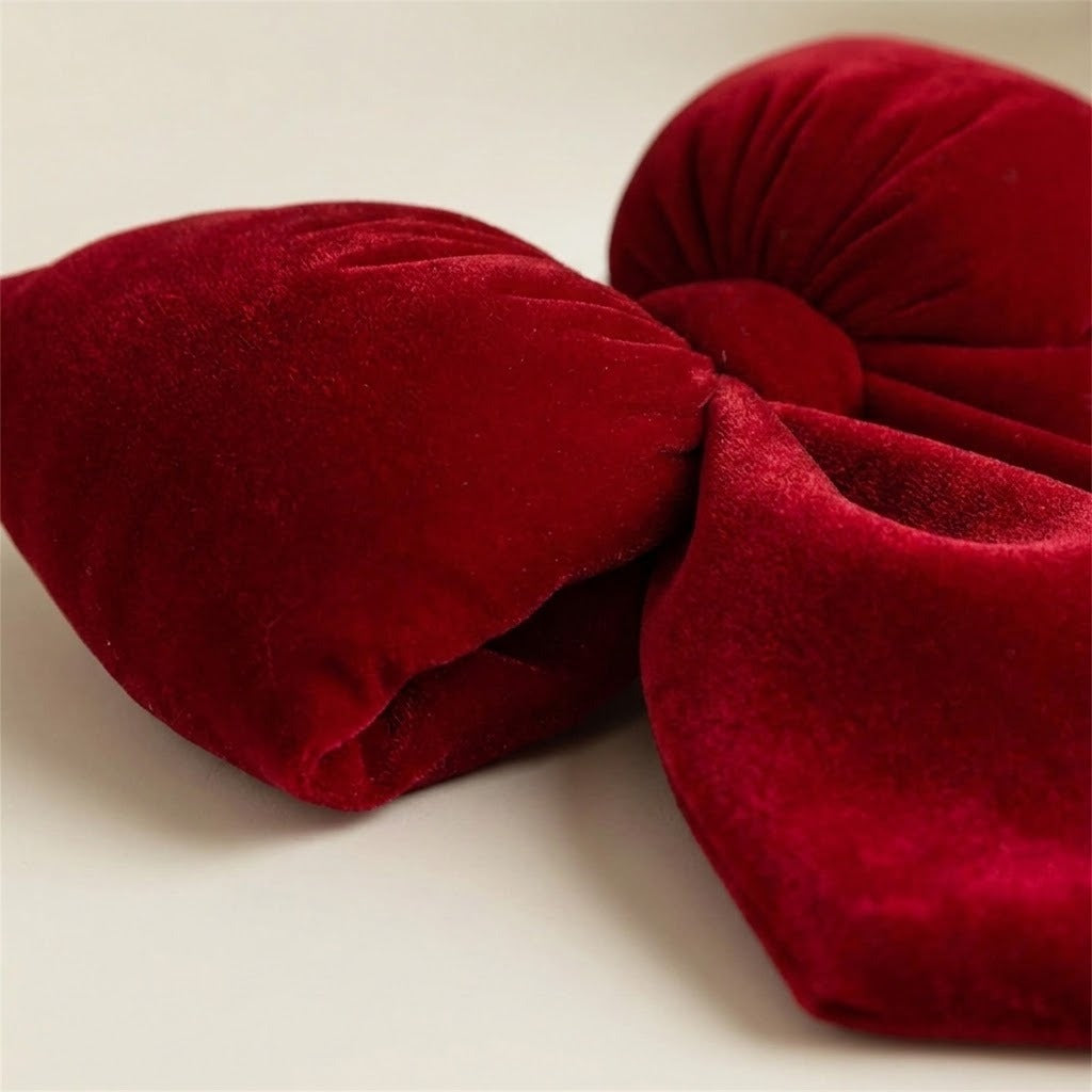 A red velvet Christmas bow rests on a cream surface, with its textured loops, wrapped center, and folded tail shown close up.