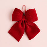 A red velvet Christmas bow with broad loops, two flared tails, and a narrow hanging ribbon fills a pale pink background.