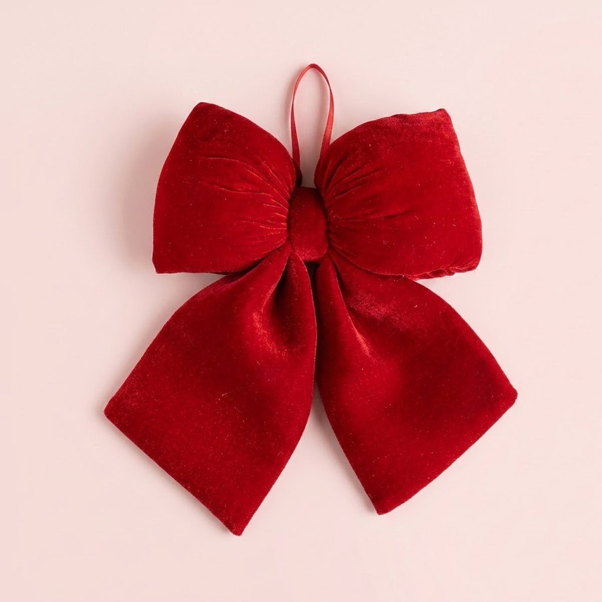 A red velvet Christmas bow with broad loops, two flared tails, and a narrow hanging ribbon fills a pale pink background.