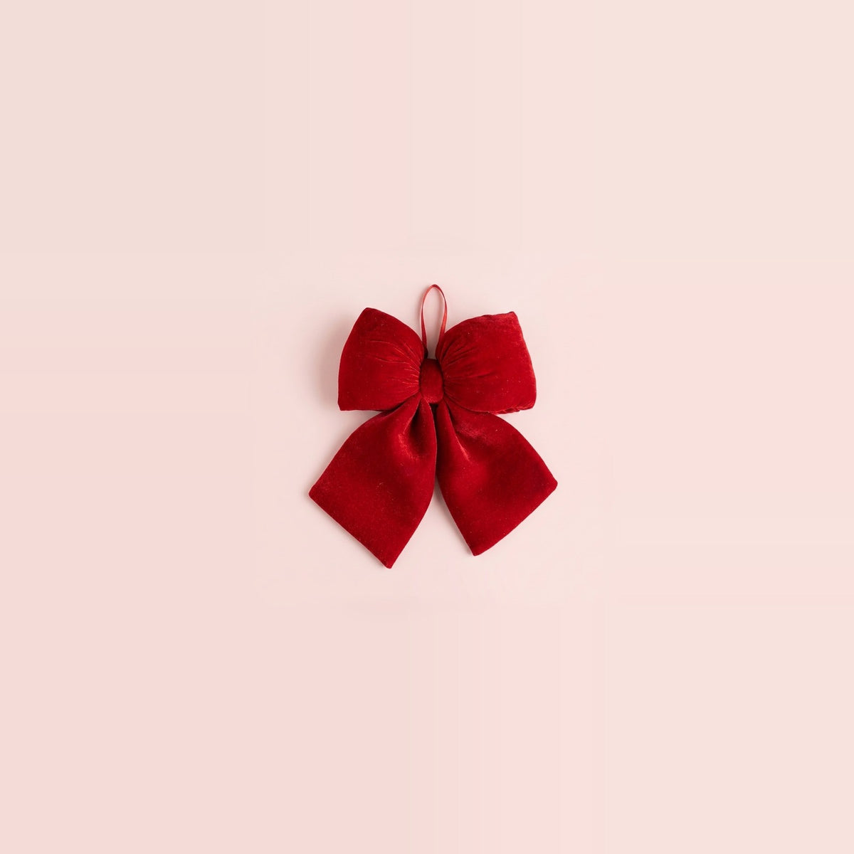 A red velvet Christmas bow with two broad tails and a ribbon hanging loop sits centered on a spacious pale pink background.