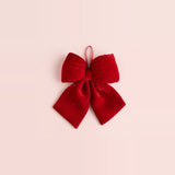 A red velvet Christmas bow with folded loops, flared tails, and a narrow hanging ribbon sits against a pale pink background.