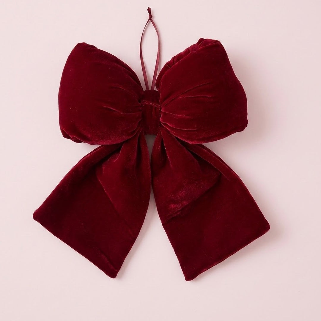 A wine-red velvet Christmas bow with gathered loops, broad tails, and a thin hanging ribbon rests against a pale pink background.