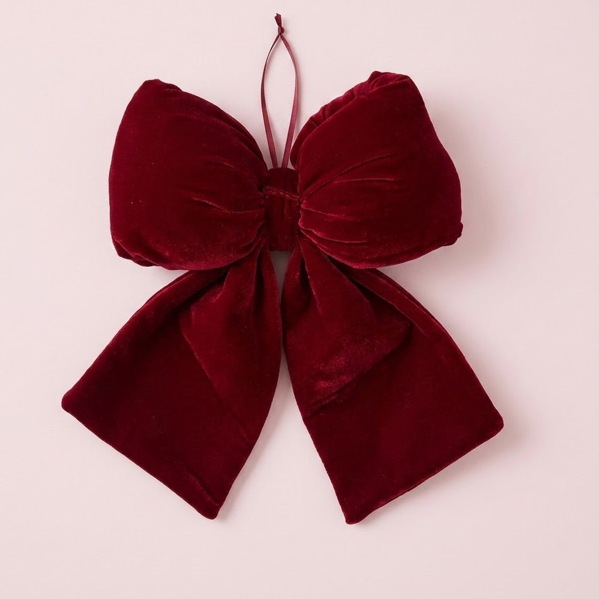 A wine-red velvet Christmas bow with gathered loops, broad tails, and a thin hanging ribbon rests against a pale pink background.
