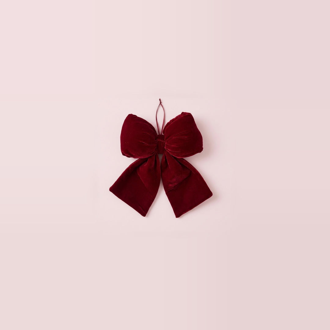 A wine-red velvet Christmas bow with gathered loops, two flared tails, and a hanging ribbon sits centered on a pale pink background.